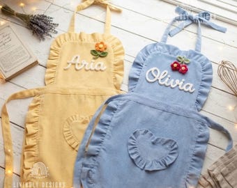 May include: Two ruffled aprons, one yellow and one blue, featuring tie-neck straps and a heart-shaped pocket. The yellow apron displays the name Aria with a crocheted flower, while the blue apron displays the name Olivia with a crocheted cherry.