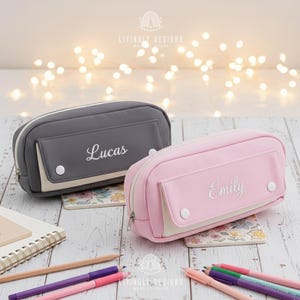 May include: Two personalized pencil cases, one gray and one pink, each with a name embroidered in white. The gray case says "Lucas," and the pink case says "Emily." Both cases have a pocket and a zipper closure. The background includes pencils and pens.