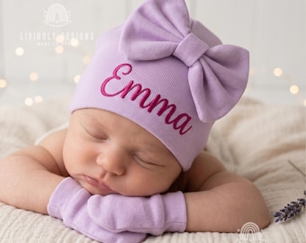 May include: A light purple baby hat featuring a large fabric bow on the front and the name Emma embroidered in dark pink script. The hat is paired with matching light purple baby mittens. The design includes the text Livinly Designs Make It Yours.