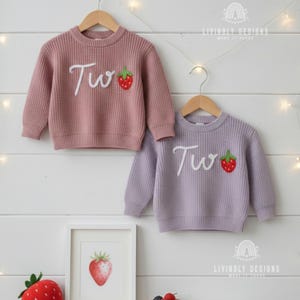 Peut inclure: Deux pulls pour enfants, un rose poudré et un lavande, sont suspendus à des cintres en bois. Chaque pull porte le mot "Two" brodé en écriture blanche, avec un motif de fraise rouge. Les pulls sont tricotés.