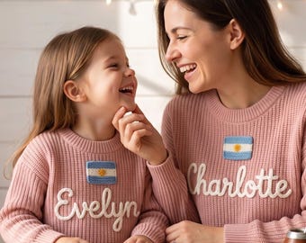 May include: Two matching dusty pink knit sweaters featuring a crew neckline and long sleeves. Each sweater displays a rectangular patch of the Argentine flag above white embroidered cursive text reading Evelyn on one and Charlotte on the other.