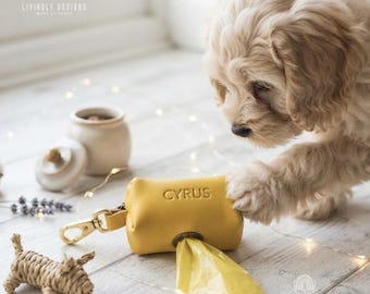 May include: A mustard yellow leather dog waste bag dispenser featuring the name CYRUS embossed on the front. The cylindrical pouch includes a gold-toned metal clasp and a circular opening at the bottom for dispensing a yellow plastic waste bag.
