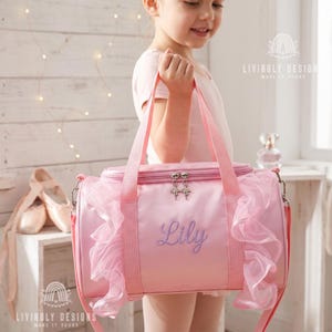 May include: A light pink cylindrical duffel bag featuring ruffled tulle trim on both sides, two top handles, and a zippered closure with ballet slipper charms. The name Lily is embroidered in purple script on the front of the bag.