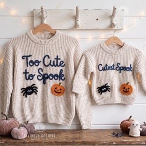 May include: Two cream-colored Halloween sweaters, one adult and one child size, hang on wooden hangers. The adult sweater says "Too Cute to Spook" and the child's says "Cutest Spook". Both have a black spider and orange pumpkin applique.