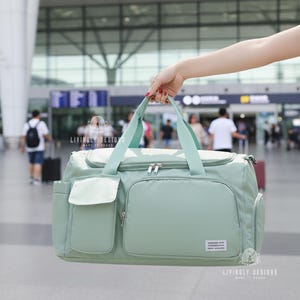 K&ouml;nnte beinhalten: Eine hellgr&uuml;ne Reisetasche mit Rei&szlig;verschluss oben, zwei Seitentaschen und einer Vordertasche. Die Tasche hat hellgr&uuml;ne Griffe und ein kleines rechteckiges Etikett auf der Vorderseite. Die Tasche wird vor einem Flughafenterminal gehalten.