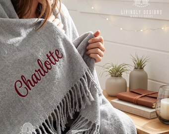 May include: A gray woven throw blanket featuring the name Charlotte embroidered in dark red script lettering. The blanket has a textured fabric finish and decorative fringe along the bottom edge. The logo Livinly Designs is visible.