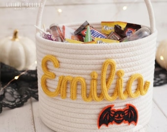 May include: A white rope basket with a handle, featuring the name Emilia in yellow cursive rope lettering and a black and orange glittery bat applique. The basket is filled with various assorted candy bars and small treats.