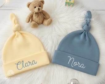 May include: Two soft baby hats with knotted tops, one in pale yellow embroidered with the name Clara and one in dusty blue embroidered with the name Nora. The hats feature a folded brim and are displayed alongside a small teddy bear.
