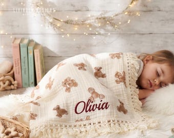 May include: A cream-colored muslin baby blanket featuring a repeating pattern of brown teddy bears and the name Olivia embroidered in dark red script. The blanket is finished with a decorative cream fringe trim along the edges.