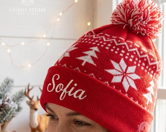 May include: A red knit beanie featuring a white snowflake and pine tree pattern, topped with a red and white pom-pom. The cuff displays the name Sofia in white script lettering. The hat is constructed from a soft, textured knit fabric.