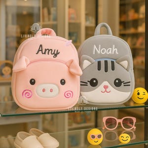Op de afbeelding: Twee kinderrugzakken, één roze varkensvormig en één grijze katvormig, elk met een naam op de voorkant. De varkensrugzak heeft de naam "Amy" en de kattenrugzak heeft de naam "Noah".