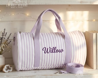 May include: A cylindrical duffel bag featuring a white and light purple vertical striped pattern. The bag includes two light purple fabric handles and a detachable shoulder strap. The name Willow is embroidered in dark purple script on the front.