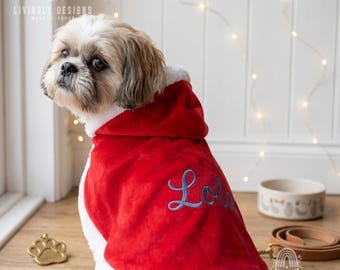 May include: A small dog with long brown and white fur wearing a red velvet hooded cape with white faux fur trim. The cape features the name Lola embroidered in blue script on the back. The garment is designed for pets and has a soft texture.