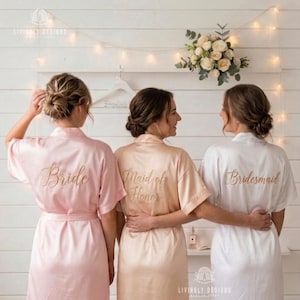 Puede incluir: Tres batas de satén en rosa claro, champán y blanco, cada una con texto bordado en dorado en la espalda. La bata de la izquierda dice Bride, la del centro Maid of Honor y la de la derecha Bridesmaid. Todas las batas tienen mangas cortas.