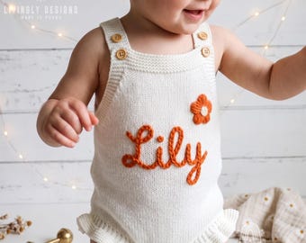 May include: A cream-colored knitted romper featuring a ruffled hem, four wooden buttons at the shoulders, and the name Lily embroidered in burnt orange yarn. A small burnt orange flower is stitched above the name on the front of the garment.