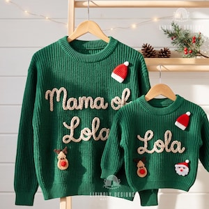 May include: Two green knit jumpers featuring cream-coloured yarn lettering. The larger jumper reads Mama of Lola, while the smaller one reads Lola. Both jumpers are decorated with small crocheted Santa hats and reindeer faces.