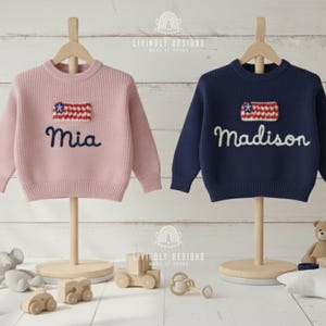 May include: Two children's sweaters, one pink and one navy blue, displayed on wooden stands. Each sweater features a flag design and a name embroidered in cursive. The pink sweater says "Mia," and the navy sweater says "Madison."