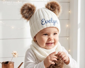 May include: A white ribbed knit beanie featuring two large brown faux fur pom-poms on top and the name Evelyn embroidered in dark blue script lettering on the front cuff. The hat is worn with a matching white ribbed knit scarf.