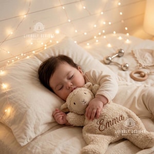 Peut inclure: Un bébé endormi, vêtu d'un pyjama crème, serre contre lui un mouton en peluche beige brodé du nom "Emma". Le bébé est allongé sur un oreiller crème. L'arrière-plan comprend des guirlandes lumineuses et un mur en bois.