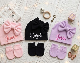 May include: Three baby hats with large bows and matching mittens in light pink, black, and light purple. The hats are personalized with the embroidered names Jenna, Hazel, and Josie in a cursive script font, arranged with baby accessories.