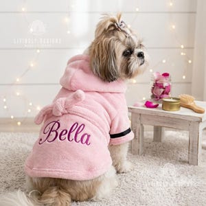 Könnte beinhalten: Ein hellrosa Plüsch-Bademantel für Hunde mit Kapuze und Bindegürtel. Auf der Rückseite ist der Name Bella in dunkelvioletter Schreibschrift eingestickt. Die Ärmel haben ein schwarzes Querstreifen-Detail an den Bündchen, der Stoff ist weich.