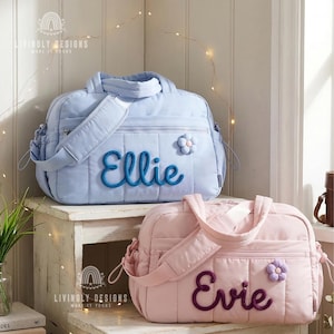 Könnte beinhalten: Zwei gesteppte Baby-Taschen, eine hellblaue und eine rosa, stehen auf einer weißen Holzoberfläche. Die blaue Tasche trägt den Namen "Ellie" in blauer Schrift, und die rosa Tasche trägt den Namen "Evie" in lila Schrift. Jede Tasche hat eine Blumenapplikation.