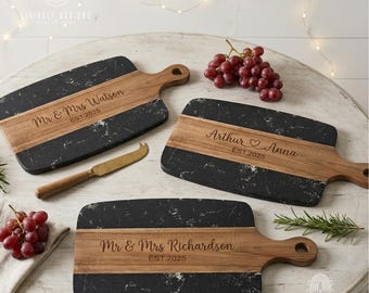 May include: Three rectangular serving boards featuring a combination of black marble with white veining and natural brown wood. Each board is engraved with names and the year 2025. A small gold-toned cheese knife with a wooden handle is included.