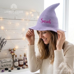 May include: A light purple knit witch hat featuring a pointed crown and a wide brim. The hat is embroidered with the name Sarah in a dark pink cursive script font on the front. The material has a fine, textured knit pattern throughout the surface.
