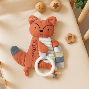May include: A plush orange fox-shaped baby toy with a white ring, holding colorful stacked blocks. The name "Lillian" is embroidered on the fox. Wooden toys, including a star and a flower, are also present.