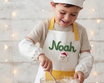 May include: A white chef hat and a white apron with yellow straps, featuring the name Noah in green embroidered script and a small Santa Claus face appliqué. The apron is worn over a beige t-shirt with white elasticized sleeve covers.