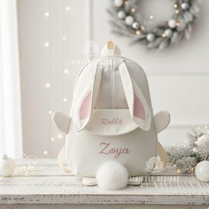 Peut inclure: Un sac à dos blanc en forme de lapin avec des oreilles intérieures roses et une queue touffue. Le sac à dos porte le nom "Zoya" brodé en script rose sous le mot "Rabbit". Il possède une poignée supérieure et des bretelles réglables.