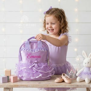 May include: A lavender backpack with a tutu-style skirt and the name "Emma" embroidered in pink. The backpack has a top handle and a bow. A young girl is smiling and holding the backpack. Other items include a stuffed bunny, shoes, and blocks.