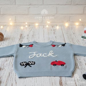 Op de afbeelding: Een lichtblauwe gebreide trui met de naam "Jack" geborduurd in wit script. De trui heeft geborduurde auto- en racethema-ontwerpen in rood, zwart en wit. De merknaam "LIVINOLY DESIGNS" is boven en onder gedrukt.