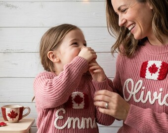May include: Two matching dusty rose knit sweaters featuring a white and red Canadian flag patch and white embroidered names. One sweater displays the name Emma, and the other displays the name Olivia. A matching ceramic mug with a red maple leaf.