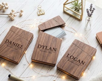 May include: Three rectangular dark wood business card holders with hinged lids. Each holder is laser-engraved with a name, DANIEL, DYLAN, or LOGAN, and the text BUSINESS CARD. The holders are arranged on a light surface with string lights.