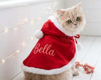 May include: A fluffy cat wearing a red hooded cape with white faux fur trim and white pom-poms. The cape features the name Bella embroidered in a script font on the back. The garment is designed with a hood and ties at the neck for a secure fit.