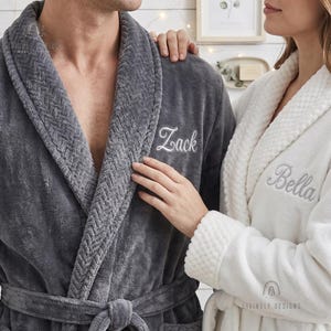 Puede incluir: Una bata de felpa gris oscuro con cuello de textura de espiga y el nombre Zack bordado, junto a una bata de felpa blanca con cuello de textura de panal y el nombre Bella bordado, ambas con mangas largas y cinturón de lazo.