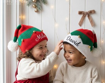 May include: Two knit elf hats featuring a red, green, and white chevron pattern with small gold bells and white pom-poms. One hat is embroidered with the name Mila in white script, and the other is embroidered with the name David in black script.