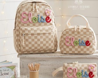 May include: A beige and white checkered backpack, lunch bag, and pencil case set. Each item features the name Eloise in colorful, raised yarn lettering and a small pink flower appliqué. The text Livinly Designs Make It Yours is visible.