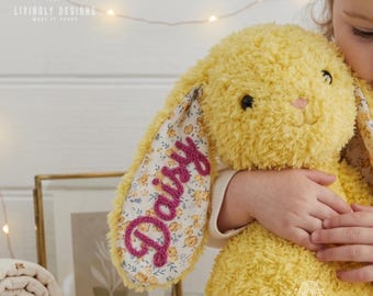 May include: A plush yellow stuffed rabbit with soft, textured fur. One ear features a floral fabric lining with the name Daisy embroidered in dark pink thread. The rabbit has black embroidered eyes and a small pink stitched nose.