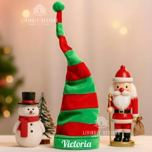 Op de afbeelding: Een vrolijke kerstscène met een lange, rood-groene elfenmuts met de naam "Victoria" op de rand. Een sneeuwpop met een rode sjaal en een kerstman notenkraker zijn ook aanwezig, wat een feestelijke sfeer oproept.