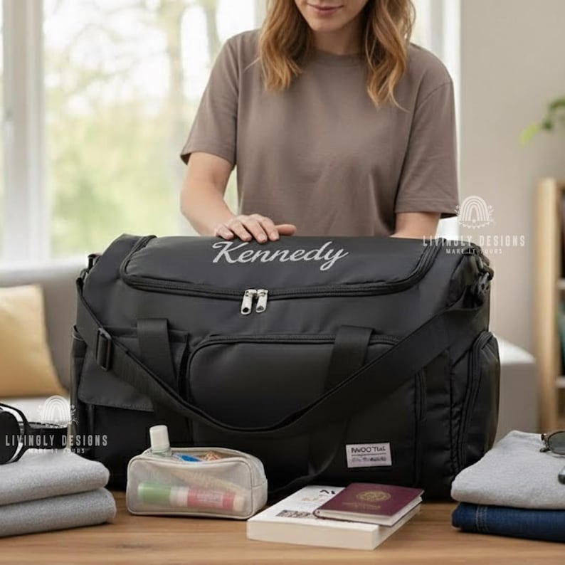 K&ouml;nnte beinhalten: Eine schwarze Reisetasche mit dem Namen "Kennedy" in Wei&szlig; gestickt. Die Tasche hat mehrere Taschen, einen Schultergurt und eine kleine transparente Kosmetiktasche mit verschiedenen Gegenst&auml;nden. Ein Reisepass und ein Buch sind ebenfalls sichtbar.
