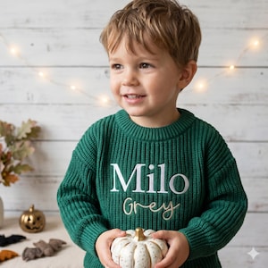 May include: A green knitted jumper with the name "Milo" embroidered in white and "Grey" in a cursive font. A child is holding a white and gold pumpkin. The background features fairy lights and autumn decorations.