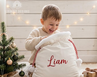 May include: A white velvet Christmas gift sack featuring a drawstring closure with red and white striped cords and pom-poms. The name Liam is embroidered in red script on the front of the sack. The sack is made of soft fabric.