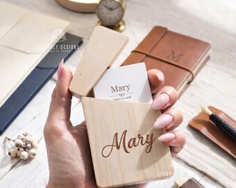May include: A light-colored wooden business card holder with the name Mary engraved in a cursive script on the front. The holder is open, revealing a white business card inside that also displays the name Mary in a simple, clean font.