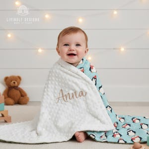 May include: A baby wrapped in a white, textured blanket embroidered with the name "Anna" in gold thread. The blanket has a blue penguin-patterned lining. The background includes a teddy bear and string lights.