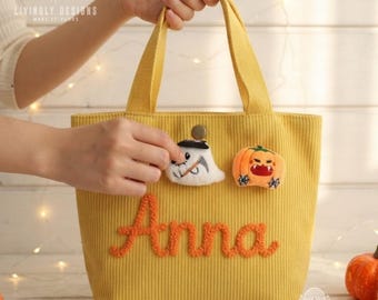 May include: A yellow corduroy tote bag featuring the name Anna embroidered in orange script. The bag is decorated with two plush patches: a white ghost wearing a black hat and an orange jack-o'-lantern with a wide, toothy grin.