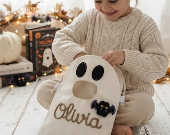 May include: A cream-colored fabric drawstring bag shaped like a ghost, featuring two black oval eyes, a round mouth opening, and the name Olivia embroidered in brown script. A small black bat applique with purple wings is attached.