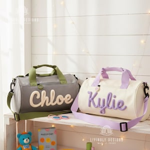 Puede incluir: Dos bolsas de lona personalizadas. Una es gris con detalles verdes y el nombre "Chloe" en escritura color crema. La otra es blanquecina con detalles lavanda y el nombre "Kylie" en escritura lavanda. Ambas bolsas tienen asas y correas para el hombro.