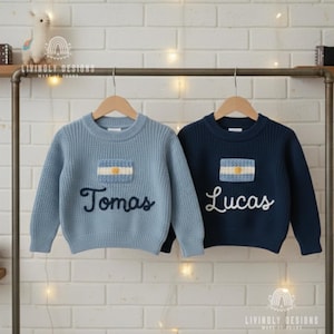 Op de afbeelding: Twee kindertruien, één lichtblauw en één marineblauw, hangen aan een houten rek. Elke trui heeft een patch van de Argentijnse vlag en een naam geborduurd in sierletters: "Tomas" en "Lucas". De truien zijn gemaakt van gebreid materiaal.